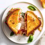 Ein mundgerechtes Chicken Parm Grilled Cheese Sandwich, umgeben von Marinara-Dip und frischem Basilikum im Sonnenlicht.  